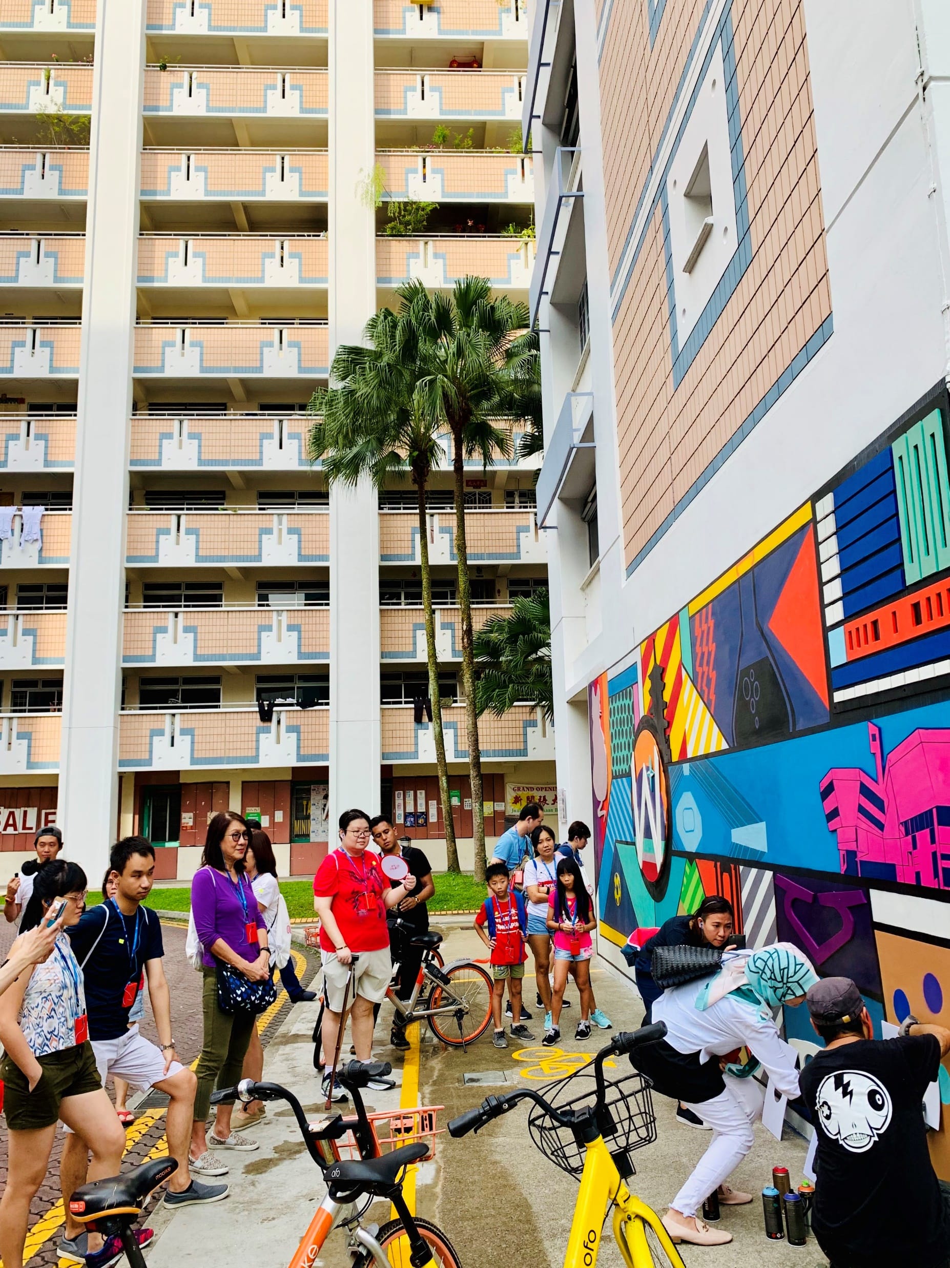People gather near a colorful mural with bicycles; in the background, there's an apartment building and palm trees.